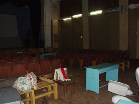 двухярусный занавес на сцене актового зала с лампами дневного света на желтых стенах, прожекторными лампами, секционными креслами старого клуба эпохи СССР