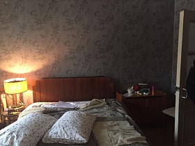 деревянная кровать с подушками на цветном постельном, прикроватная тумбочка с косметикой у стены с цветастыми обоями спальной комнаты кв.24
