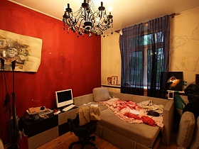 люстра , стилизованная под свечи на потолке спальной комнаты с разложенным диваном,телевизором на тумбочке и торшером у красной стены спальной комнаты разноплановой простой квартиры
