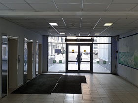 просторный светлый холл с квадратной плиткой на полу, дорожками у лифтов, цветной картиной на боковой стене и входными стеклянными дверьми в учреждение