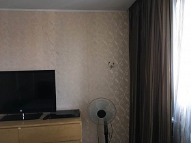 плоский черный телевизор на бежевом комоде, вентилятор у стены спальной комнаты с коричневыми шторами на окне