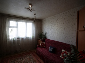 разноцветные подушечки на диване с вишневым покрывалом, комнатные цветы на комоде у большого окна с белой прозрачной гардиной светлой комнаты с белой люстрой на потолке