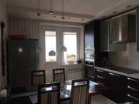 стулья вокруг длинного обеденного стола, серый холодильник и темная мебельная стенка на кухне с полосатыми обоями семейной трехкомнатной квартиры