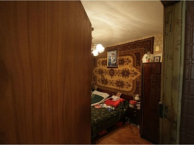две деревянные односпальные кровати с зеленым марселевым покрывалом и зеленым одеялом у стены с большим желто-коричневым ковром, портретом в рамке на стене спальной комнаты через открытую дверь квартиры бабушки и деда в стиле 80-89 гг