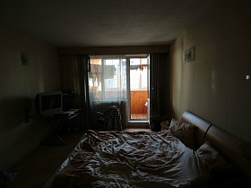 телевизор на ножке ,столик, рубашка на компьютерном стуле у окна спальни с открытой дверью на застекленный балкон трехкомнатной квартиры геолога в высотном доме