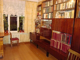уютная чистая комната с многочисленными книгами на открытых полках и полках мебельной стенки, комнатными цветами на подоконнике окна с короткой белой гардиной и желтыми шторами квартиры СССР бабушки
