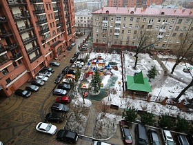 припаркованные машины вдоль дороги и жилых домов во дворе с зелеными елями, дорожками, детскими площадками за забором в зимнее время