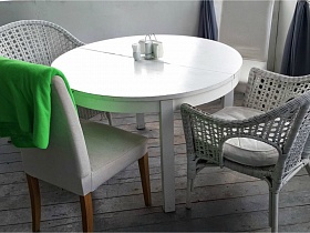 два белых плетеных кресла и мягкий стул вокруг белого деревянного стола в уголке белой веранды уютного кафе с большими окнами