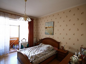 цветочная подвесная люстра на белом потолке над большой кроватью со светлым постельным, напольная вешалка для одежды у окна с белой гардиной и коричневыми шторами большой семейной квартиры