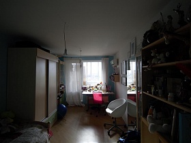 шкаф с мягкими игрушками и папками на открытых полках, большое зеркало на стене, белое кресло и письменный стол у окна с белой гардиной в спальне с голубой стеной стильной трехкомнатной квартиры