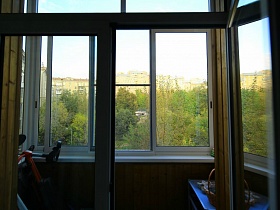 вид на зеленый участок придомовой территории многоэтажных жилых домов с окна застекленного балкона семейной трешки
