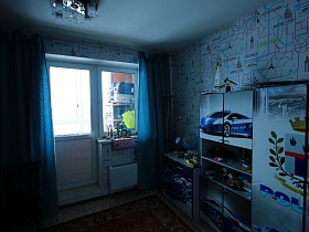 голубая мебель с машинами на дверцах шкафов, голубая кровать в виде машины, голубые шторы на окне с балконной дверью детской спальной комнаты простой современной квартиры