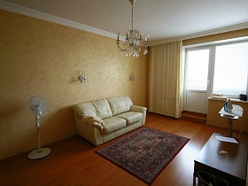вентилятор, цветной ковер у белого мягкого дивана с подушками в гостиной с желтыми стенами и желтыми шторами на окне  квартиры в переезде (въезде) молодоженов