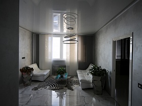 необычной формы высокие серые вазоны с комнатными цветами у белого дивана и кресла, низкий стеклянный цветной столик на коврике, стилизованном под шкуру зебры в гостиной в серых тонах с большим окном