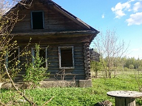 старый заброшенный деревянный дом под треугольной крышей с окнами без стекол на заросшем участке в старой деревне