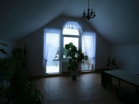 необычное окно с прозрачной белой гардиной, многочисленные комнатные цветы в вазонах на полу и полках, длинный прямоуголный стол на мансарде двухэтажного гостевого дома с придорожным кафе