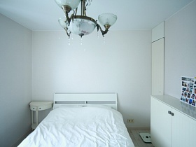 прикроватная тумбочка у большой белой кровати с белым покрывалом, напольные весы у белого шкафа с коллажем из фотографий в белой спальной комнате стильной квартиры блогера