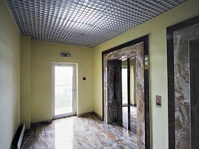 просторный коридор с желтыми стенами, мраморной плиткой на полу и вокруг лифтовых кабин в современном элитном многоэтажном доме