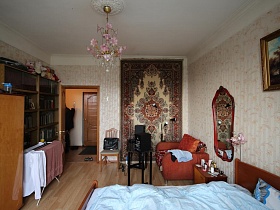 открытая дверь в спальную комнату с большим ковром на стене и розовой люстрой на белом потолке квартиры сталинки