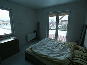 коричневый комод у окна, отопительная батарея у стены и большая кровать с цветным постельным в светлой спальне недостроенного элитного дома