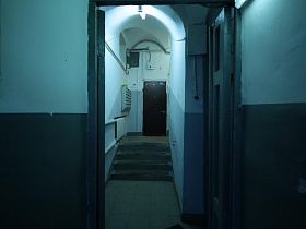коричневая дверь жилой квартиры на первом этаже серого стильного подъезда с квадратной плиткой на полу перед ступенями лестницы с перилами