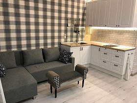 серый мягкий угловой диван с подушками у стены с клетчатыми обоями, двухцветная светлая мебельная стенка с плиткой на рабочей поверхности в зоне кухни стильной дизайнерской студии однокомнатной квартиры