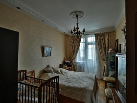 бежевые шторы с цветами и белая гардина на окне с балконной дверью в спальне с большой кроватью и детской кроваткой в трехкомнатной семейной квартире эпохи СССР