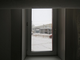 заснеженный двор с машинами на проезжей дороге, одноэтажными постройками через стекло окна квартиры на первом этаже двухкомнатной квартиры
