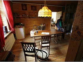 картины и деревянные полки на стене, холодильник  у окна с розовыми шторами и обеденный стол в зоне кухни деревянной дачи музыканта