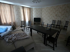 коричневые шторы на окнах, множество стульев вокруг коричневого стола, разложенный угловой диван с подушками  в просторной гостиной кирпичного двухэтажного дома для съема