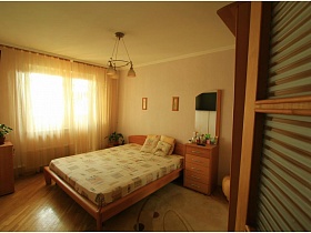зеркало с комодом,картины над большой деревянной кроватью , комнатные цветы на шкафчиках в бежевой спальне трехкомнатной квартиры