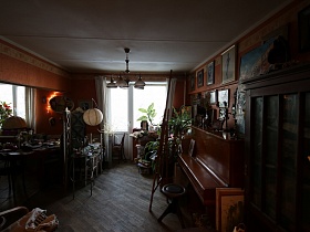 коричневый шкаф со стеклянными дверцами, круглый стул и пианино у стены с многочисленными картинами в рабочей комнате с окном и балконной дверью квартиры эпохи СССР