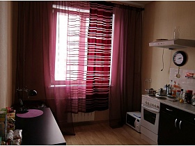 коричневая настольная лампа на сиреневой салфетке, многочисленные коробочки на поверхности обеденного стола у стены кухни с сиреневыми и фиолетовыми гардиной и шторами на окне
