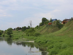 ряд домов с участками за забором, расположенных на зеленом крутом берегу красивой реки в живописном месте