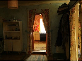 белый шкаф с открытыми полками, стол, навесная вешалка с одеждой, старые коврики на полу комнаты с цветными шторами на дверных проемах в жилом доме на селе