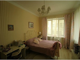 стул у письменного стола молочного цвета, комод, кровать с металлической спинкой у светлых полосатых стен спальной комнаты во французском стиле с люстрой на белом потолке