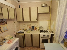 бежевая с коричневыми полосами мебельная кухня,газовая плита, белая стиральная машинка с туалетными принадлежнстями, обеденный стол у окна прихламленной квартиры бабушки и деда эпохи СССР
