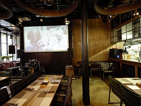музыкальные инструменты и большие колонки на сцене у декоративной коричневой стены с большим экраном в помещении крафтового ресторана с большими окнами