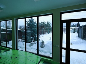 заснеженный участок с зелеными елями сквозь большие панорамные окна ярко зеленой кухни дома с частичным недостроем