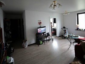 плоский телевизор на швейной машинке и небольшой столик у стены возле открытой двери и спортвный тренажер в углу гостиной простой трехкомнатной разрисованной квартиры