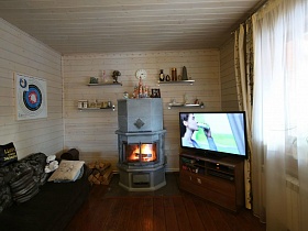 вязанка дров за диваном  у серебристого камина, телевизор на тумбе в углу комнаты, полочки на стене гостиной современного деревянного дома