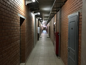 двери жилых квартир в стенах из красного кирпича длинного коридора с квадратной серой плиткой на полу