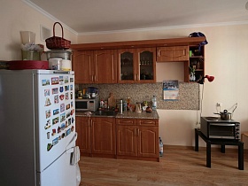 белый холодильник с магнитиками, коричневая кухня с посудой на пестрой столешнице и пестрой рабочей стенкой в простой квартире семьи в зеленом массиве , с видом на море