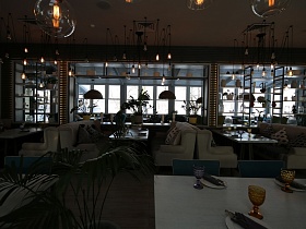 потолочные люстры в стиле хай-тек над зонами отдыха с мягкими белыми диванами у сервированных столиков в прозрачном стильном ресторане