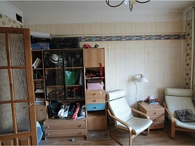 мебельная стенка с разнообразными вещами и предметами на открытых полках, два белых кресла у бежевого комода в комнате с полосатыми обоями простой сталинки 90-ых