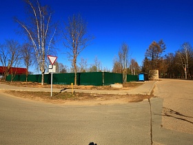 высокий зеленый забор вокруг участка с жилым домом на углу перекрестка дорог с дорожным знаком и синим павильоном на остановке в Сычево для съемок кино