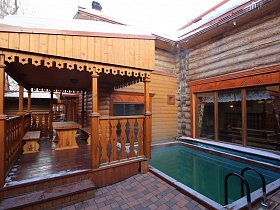 бассейн с лестницей под большими окнами деревянного дома с парной и у открытой веранды под крышей