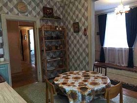 трехрошковая деревянная люстра со стеклянными плафонами на светлом потолке гостиной с прозрачной гардиной и синими шторами на окне из открытой двери кухни с круглым обеденным столом посередине