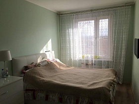 большая гардина на всю стену окна бирюзовой спальни с белой кроватью и белым комодом современной двухкомнатной квартиры