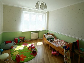 многочисленные игрушки на полу, зеленом цветном коврике,деревянной кроватке в светлой детской комнате с салатовыми панелями милой женской современной квартиры в новострое